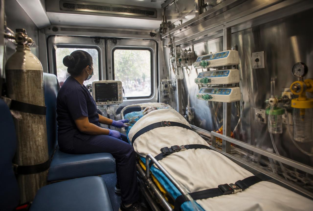 Indian critical-care nurse monitoring a patient on a secured stretcher inside an ICU ambulance with infusion pumps and oxygen.
