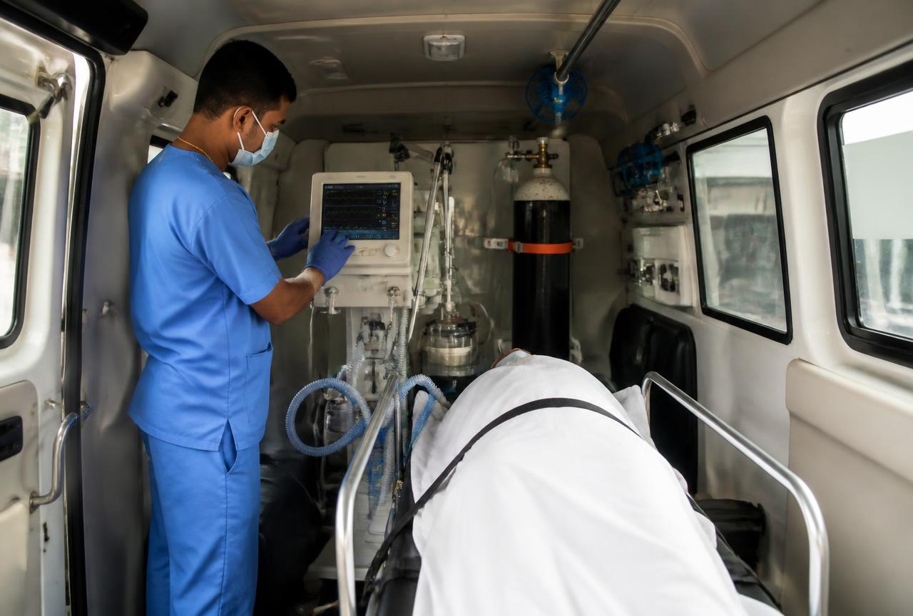 Indian respiratory therapist adjusting a transport ventilator beside a secured stretcher inside a ventilator ambulance.