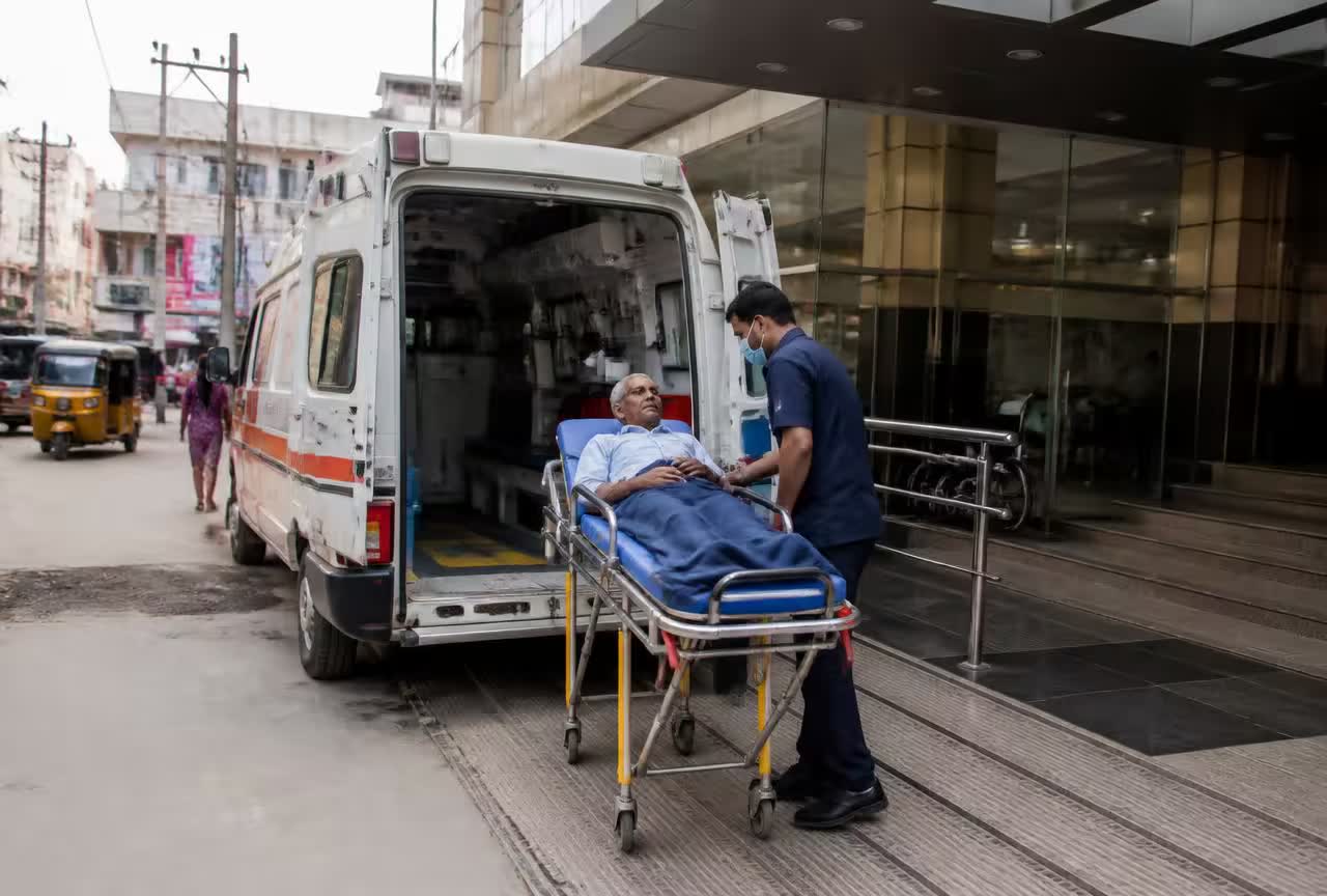 Basic Life Support ambulance parked outside a Hyderabad hospital while an Indian paramedic wheels a stable elderly patient on a stretcher trolley.